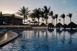 Zona de piscina con sillas de descanso y sombrillas, rodeada de palmeras y un edificio de diseño moderno.