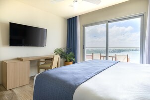 A hotel room with a bed, a desk, a TV, a potted plant, and a balcony with a view of the city.