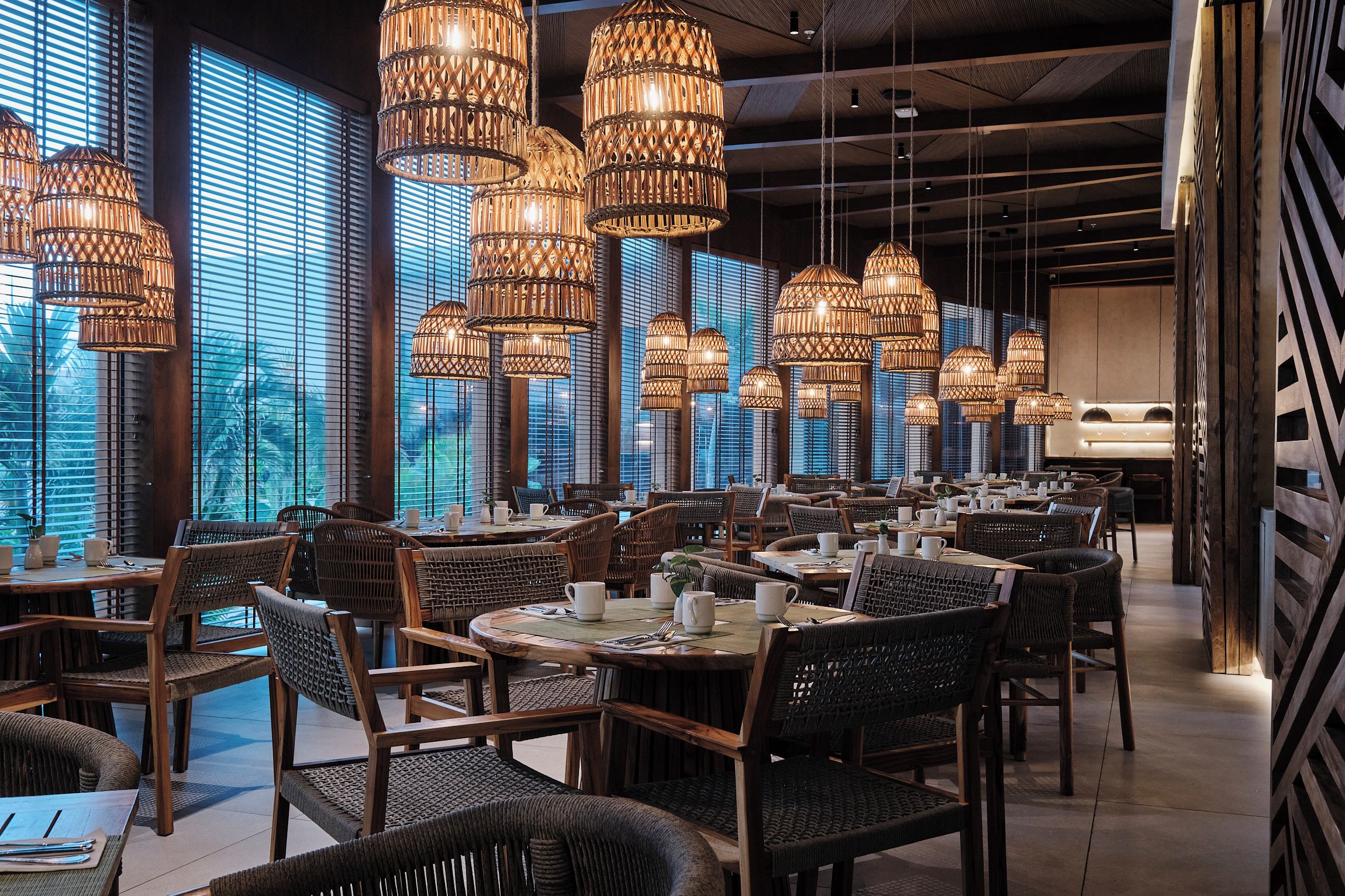 Interior de un restaurante con varias mesas y sillas, ventanas amplias con persianas y candelabros de mimbre colgantes.