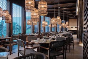 A restaurant interior with multiple dining tables and chairs, large windows with blinds, and hanging wicker chandeliers. - Hotel NYX Cancun All Inclusive (Cancun)