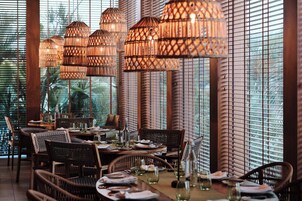 Interior de un restaurante con mobiliario de mimbre, lámparas colgantes de mimbre y vistas a través de grandes ventanales.