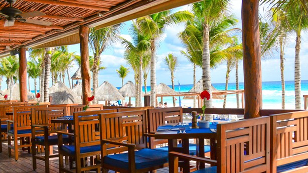 A covered outdoor seating area with wooden tables and chairs, overlooking a beach with palm trees and thatched umbrellas. - Hotel NYX Cancun All Inclusive (Cancun)