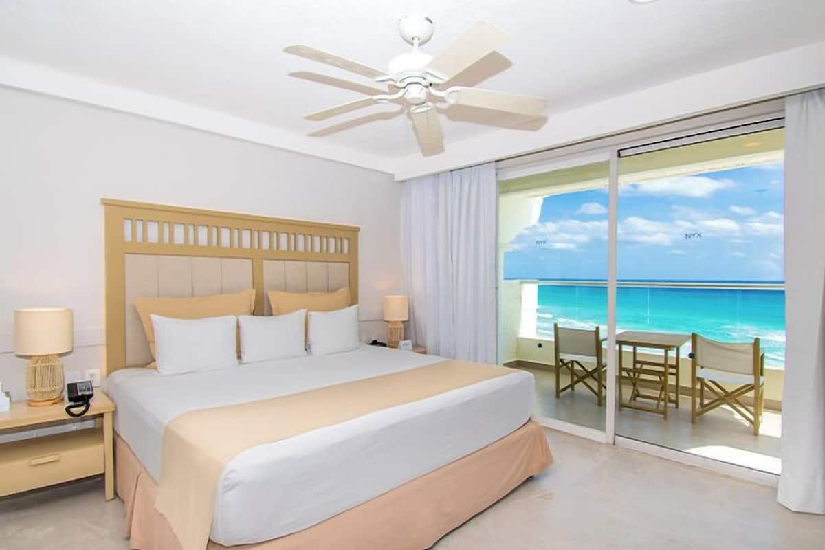 Habitación de hotel con cama, ventilador de techo, mesita de noche, lámpara, vista al océano y balcón con sillas.