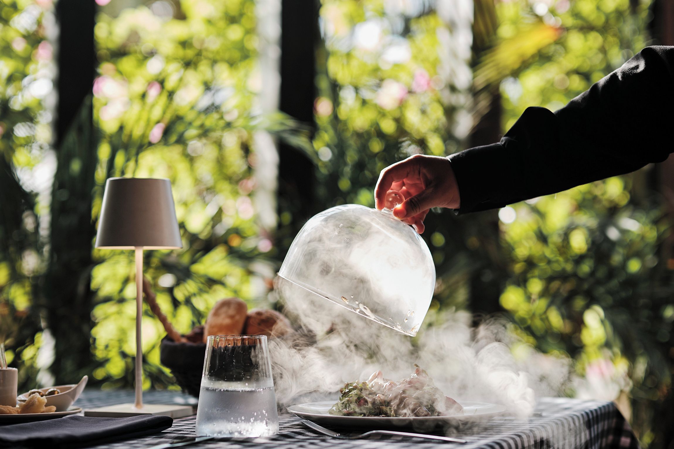A dining table with a clear glass dome over a dish, a lamp, and a glass of water.