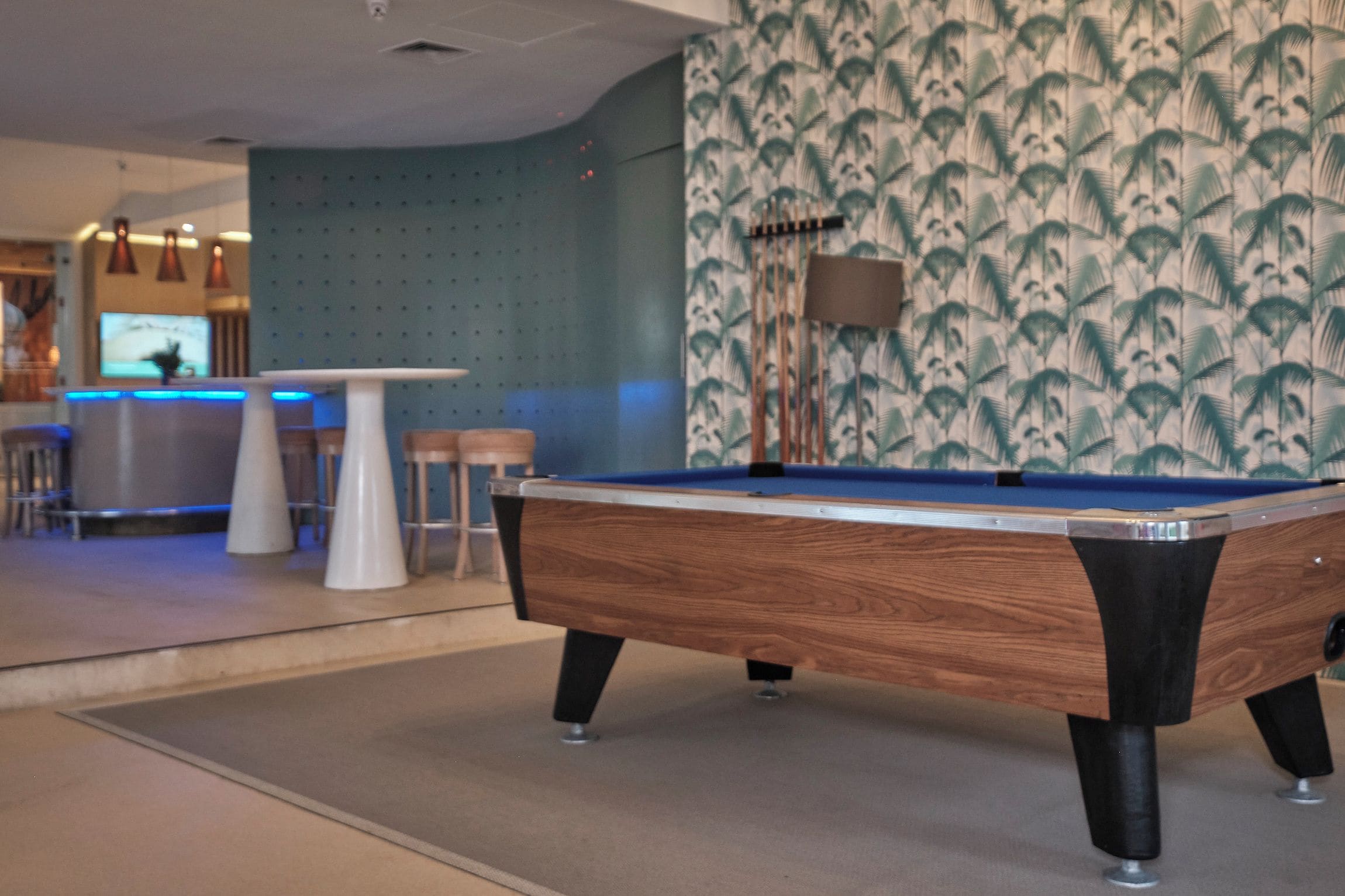 A pool table in a room with a blue wall and a patterned wallpaper. There is a bar area with stools and a television in the background.