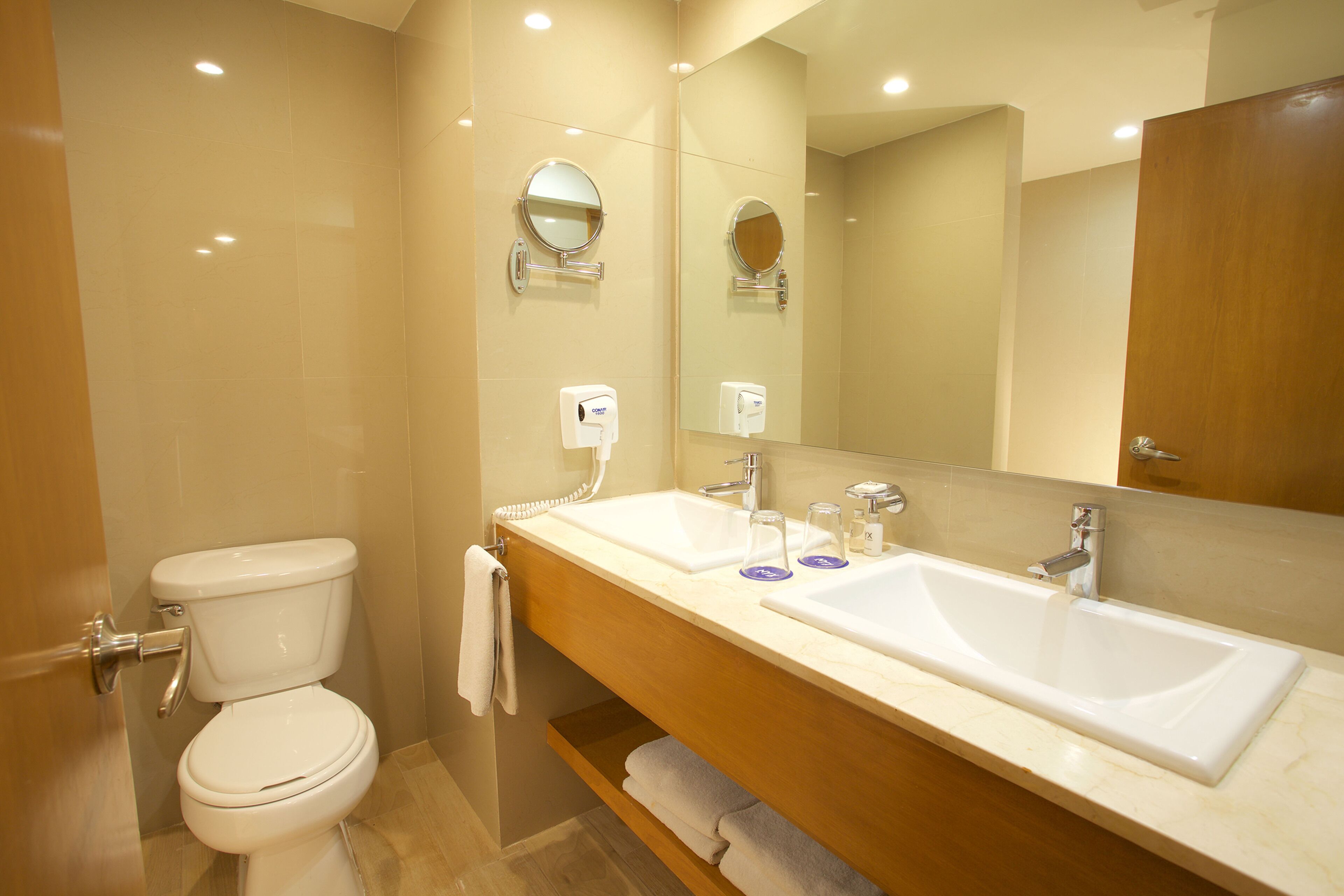 Un baño moderno con inodoro, dos lavamanos, un espejo grande y un perchero para toallas.