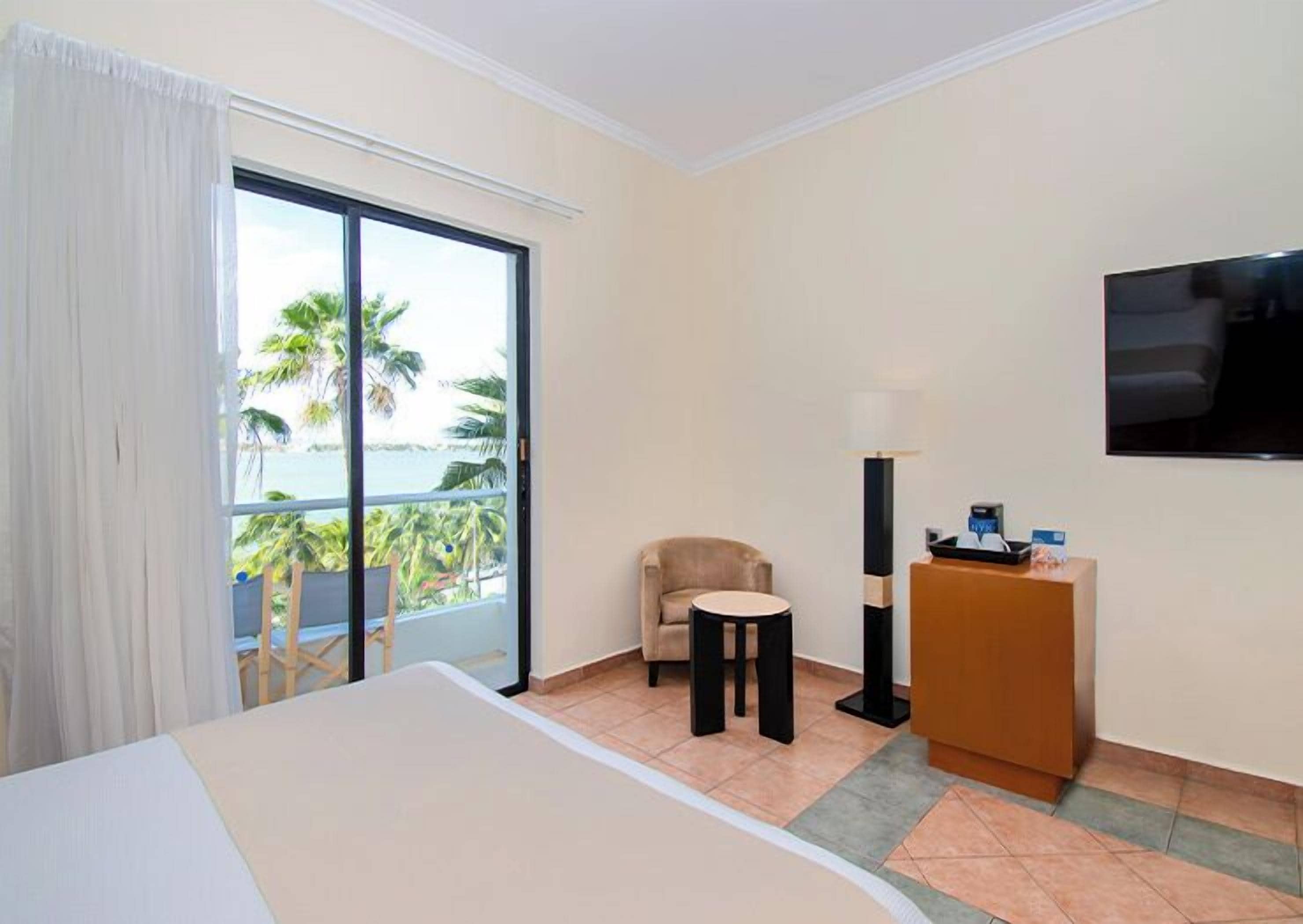 A hotel room with a bed, a chair, a desk, a TV, and a view of palm trees and the ocean.