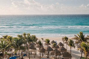 A beach with thatched umbrellas and palm trees, a sandy shore, and the ocean in the background. - Hotel NYX Cancun All Inclusive (Cancun)