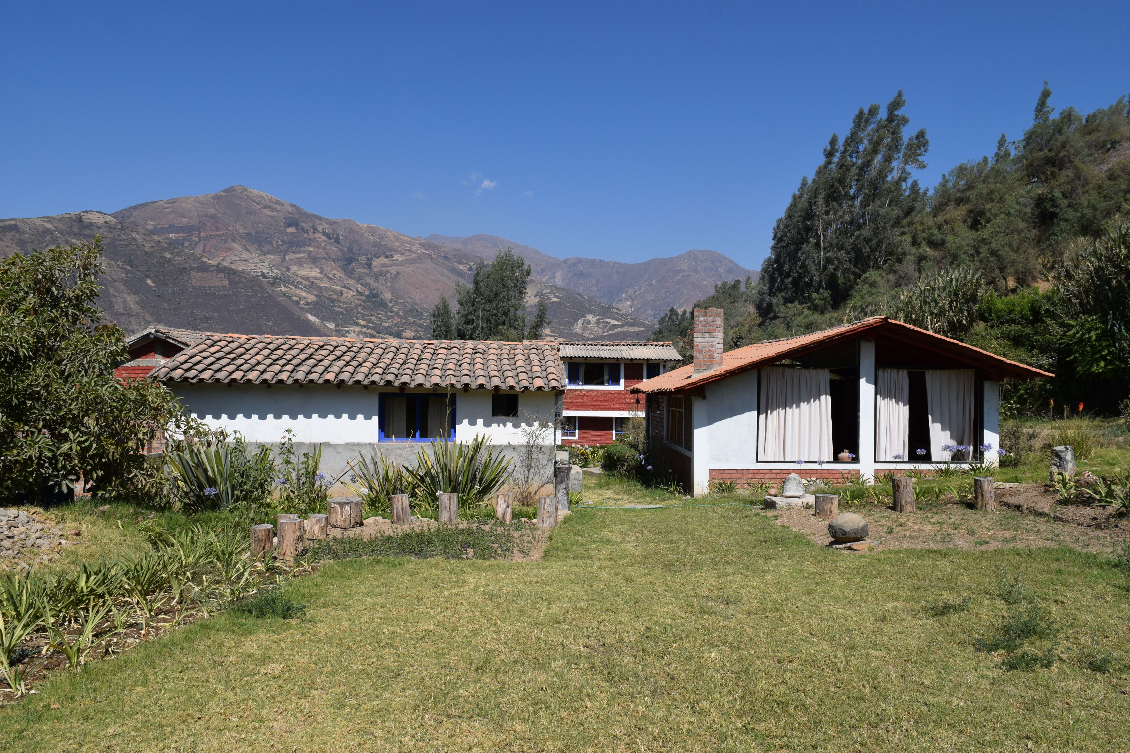 A rural house with a tiled roof, surrounded by a garden with various plants and a mountain in the background.