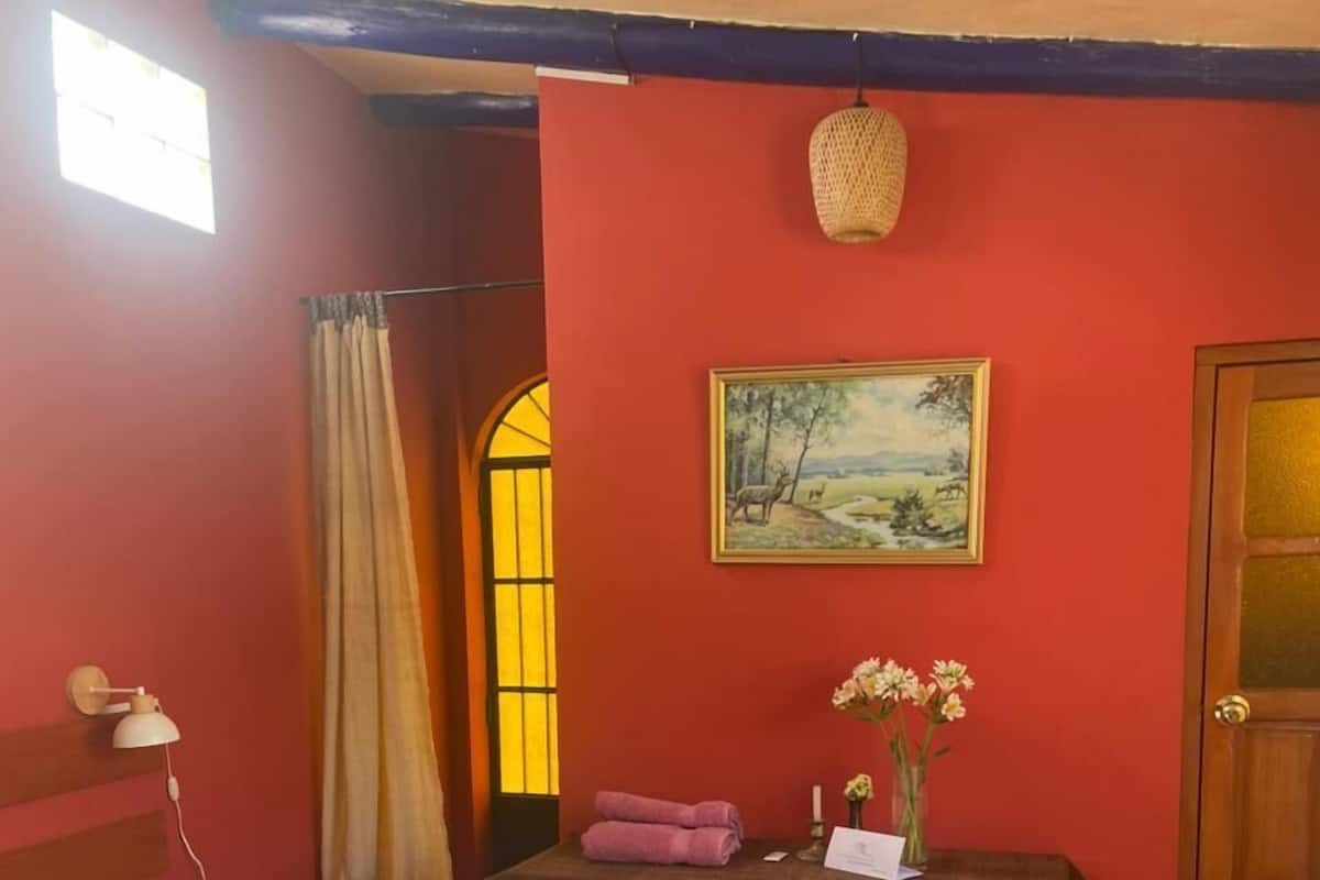 A room with two beds, a wooden floor, a painting on the wall, a table with a vase of flowers, and a door.