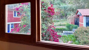 Une vue à travers une fenêtre à cadre en bois, donnant sur un bâtiment en briques rouges avec des fenêtres bleues et des fleurs roses.