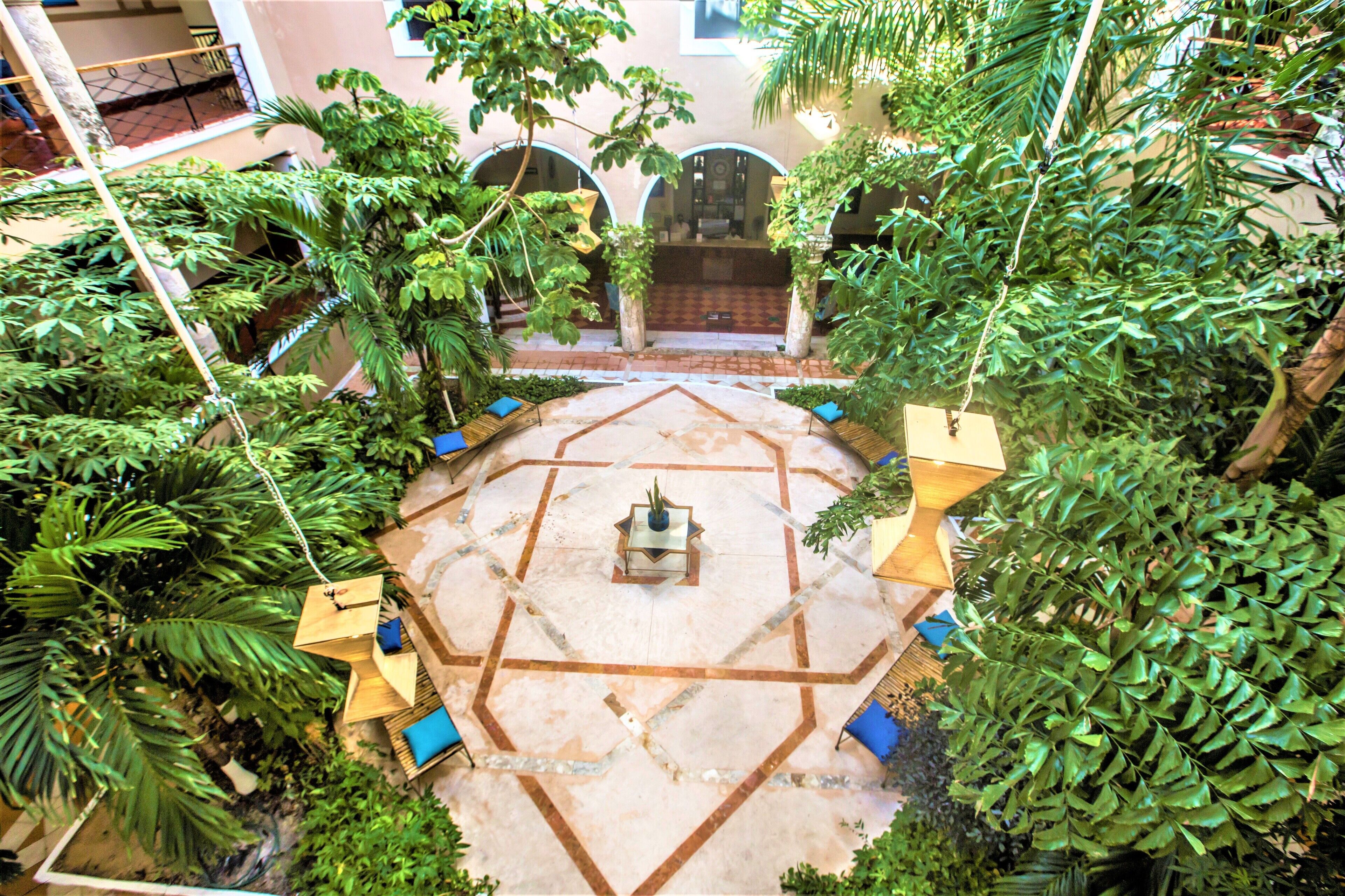 Un patio con un diseño geométrico de azulejos, rodeado de abundante vegetación y una fuente central.