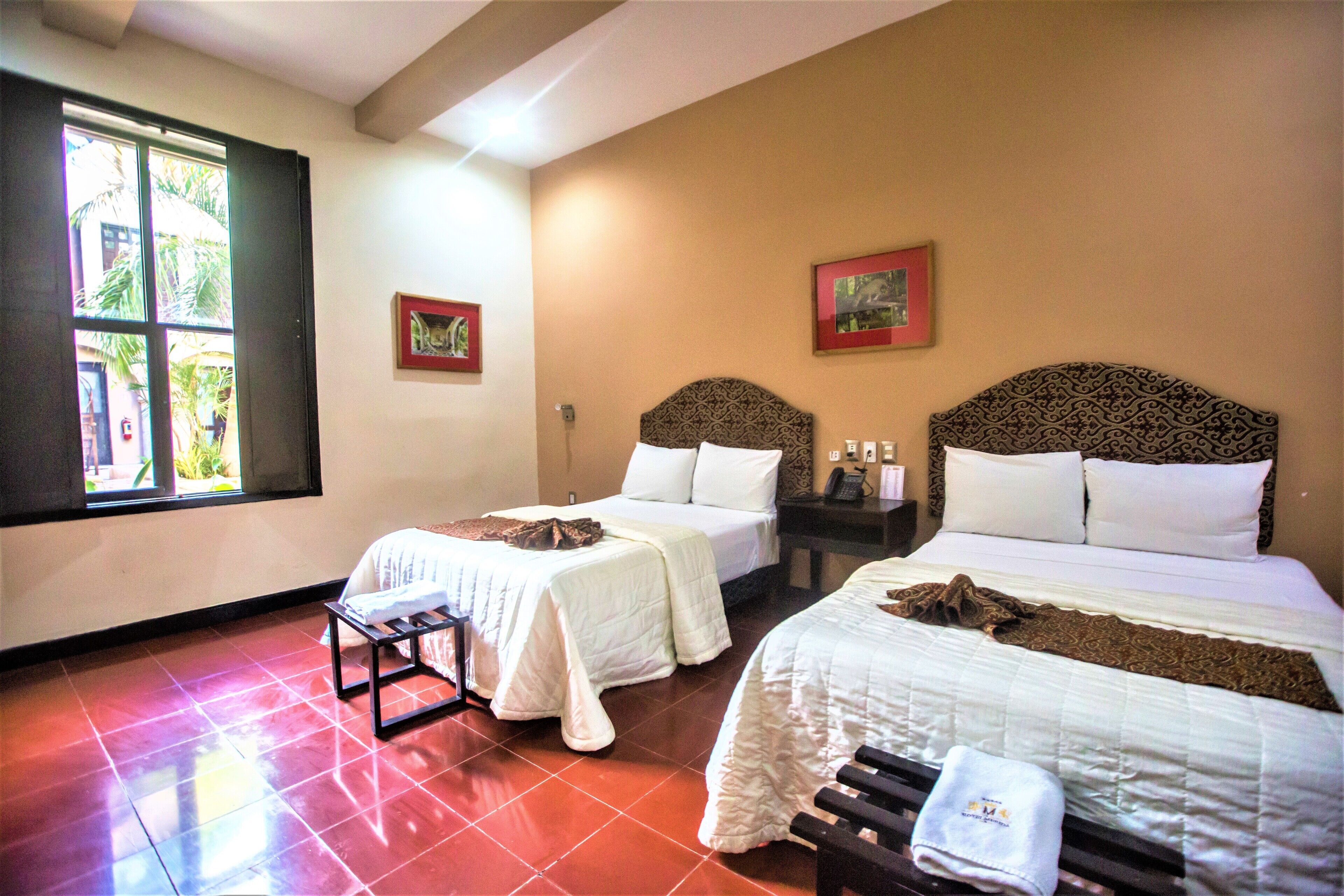 A hotel room with two beds, a small table, and a window with a view of palm trees.