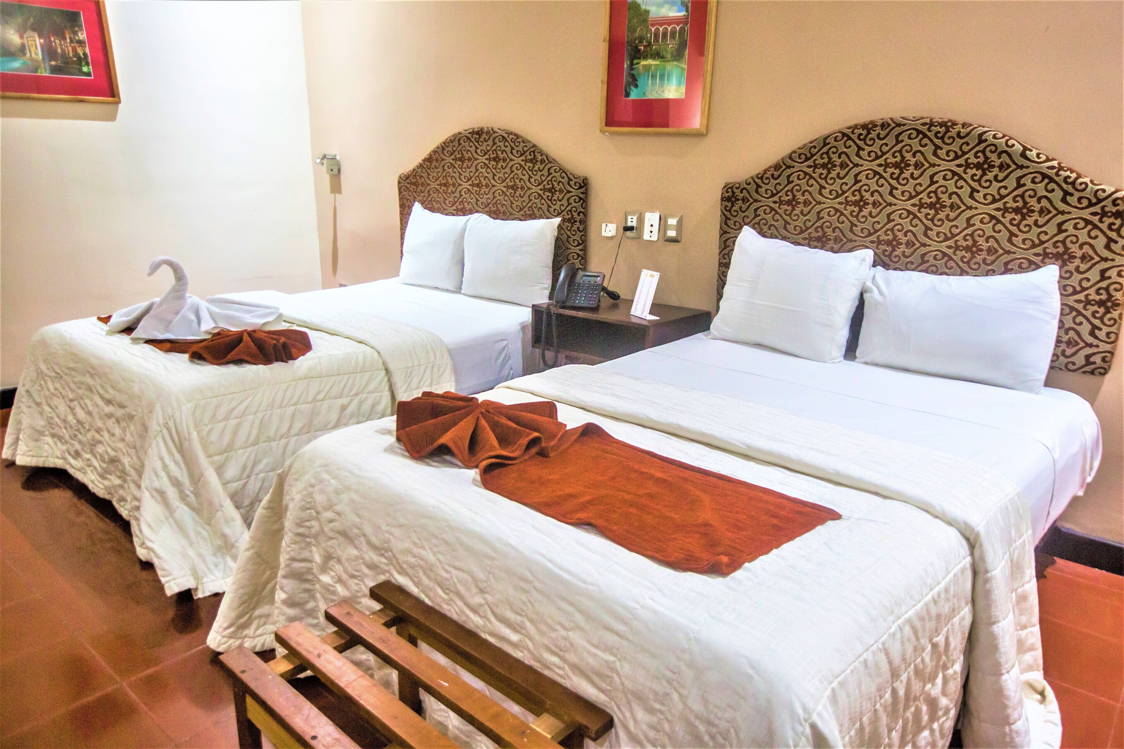A hotel room with two beds, each with a headboard, a bedside table with a telephone, and a wall-mounted light switch.