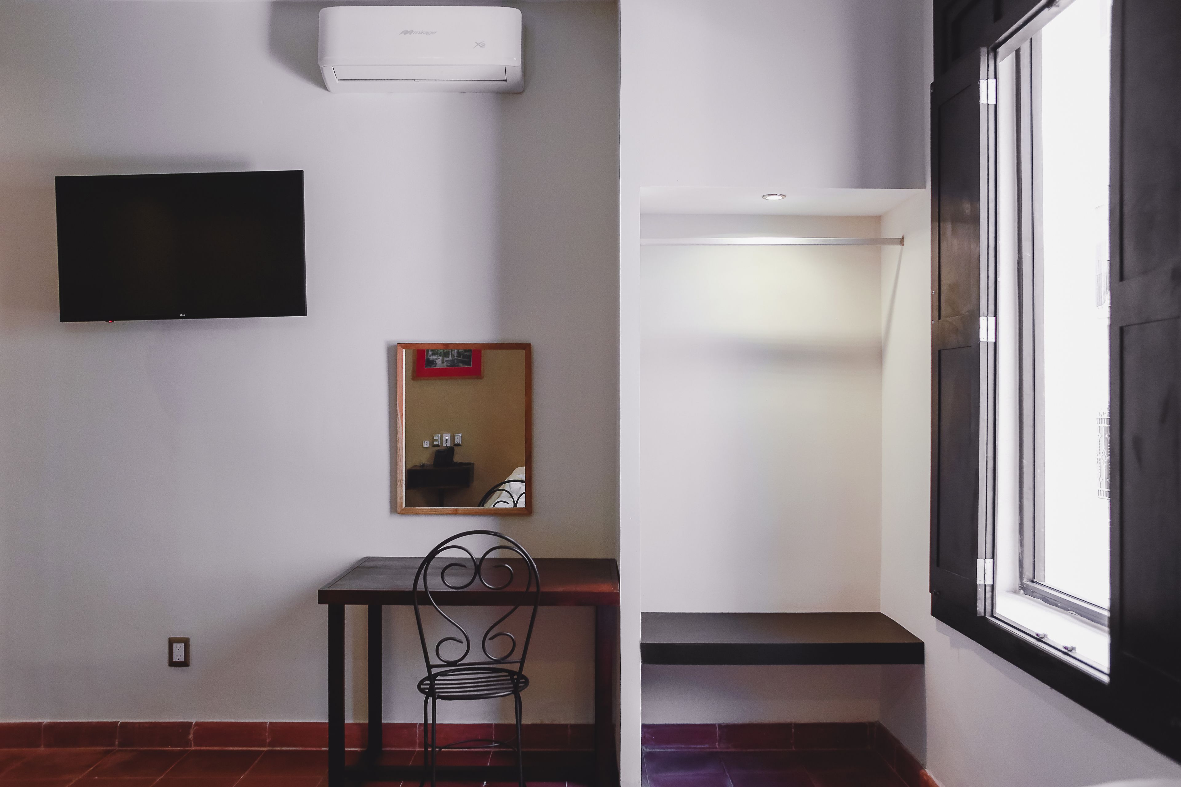 A room with a flat-screen TV mounted on the wall, a small table with a chair, a mirror, and a window with a view of the outside.