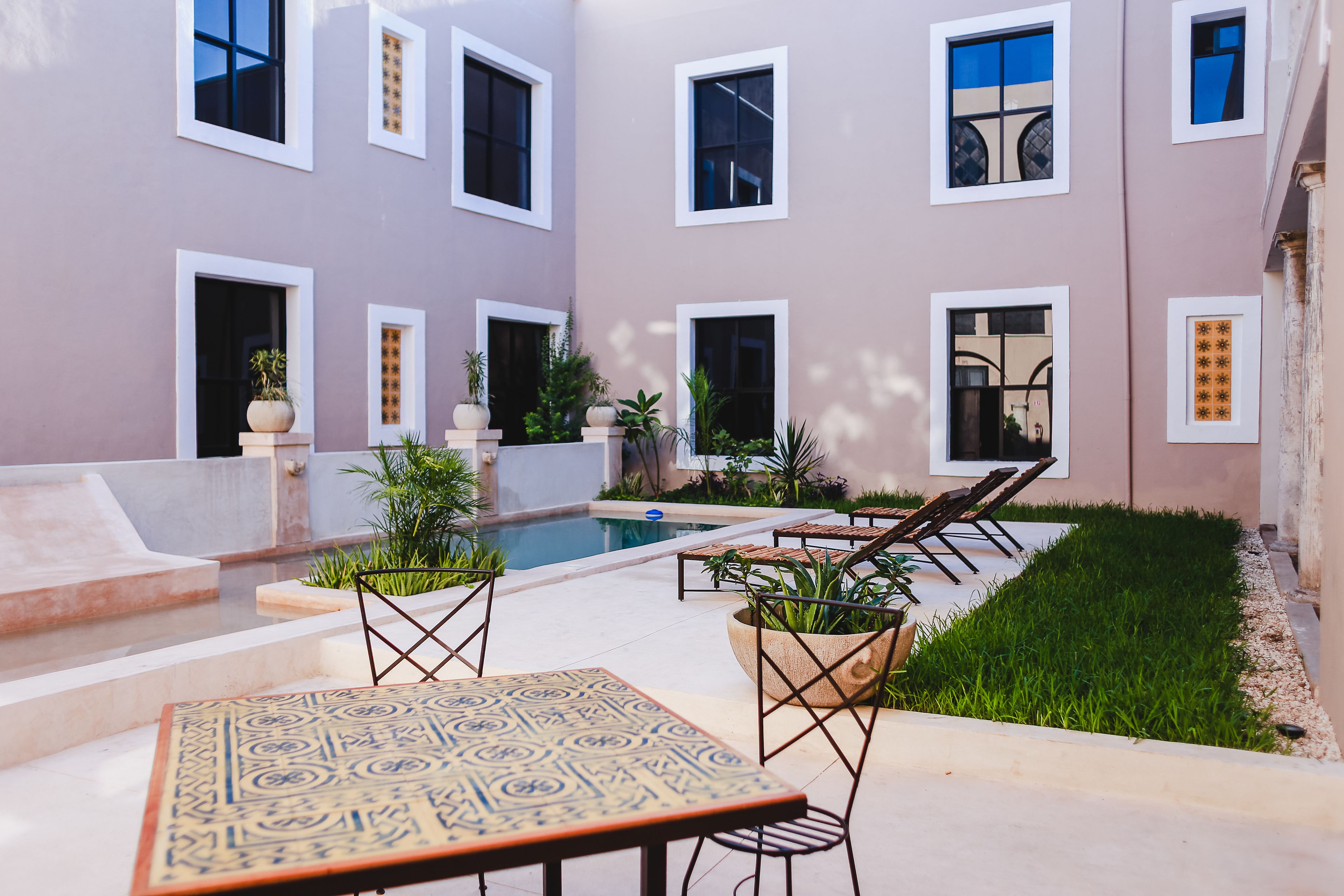 Zona junto a la piscina con mesa y sillas, rodeada de plantas en macetas y una piscina.