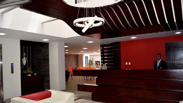 A modern reception area with a red wall, a white sofa, and a wooden reception desk. - Hotel Avanty (Ipiales)