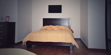A bedroom with a bed, a flat-screen TV mounted on the wall, and a wooden dresser.