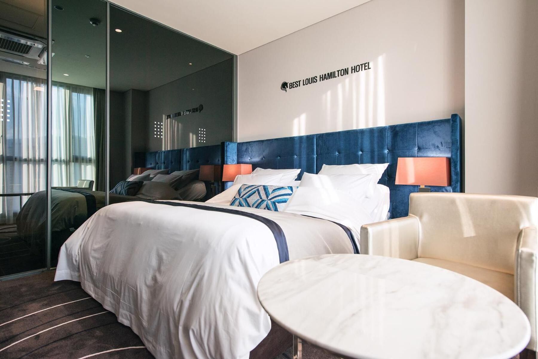 Una habitación de hotel con una cama grande, una mesa redonda y una silla. Se observa una pared con un cartel que dice "Best Louis Hamilton Hotel".