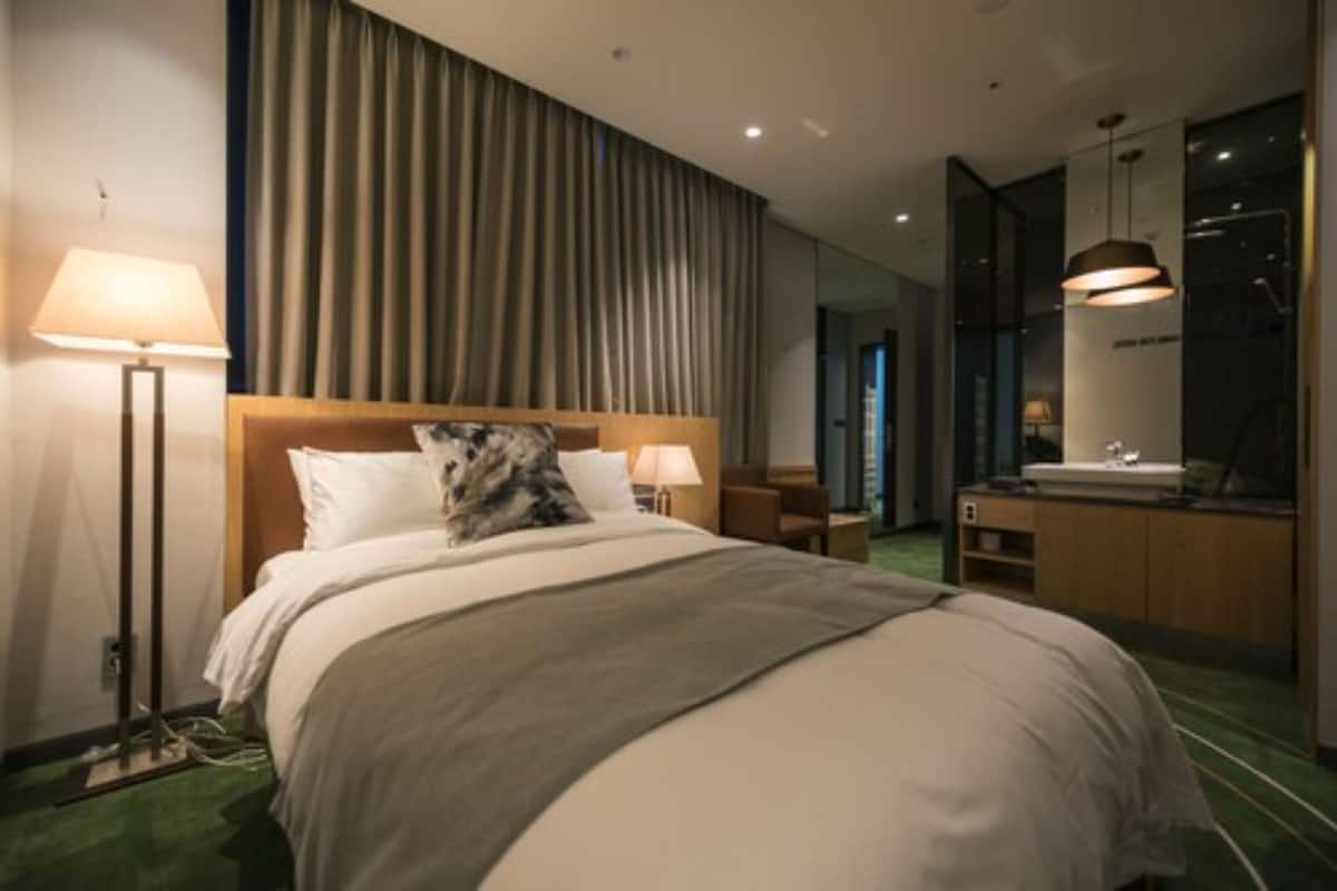 A hotel room with a large bed, a bedside table with a lamp, a bathroom with a mirror and sink, and a view of the city.