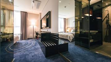 Habitación de hotel con cama, escritorio, silla, sofá, televisor, espejo y baño con ducha y mampara de vidrio.