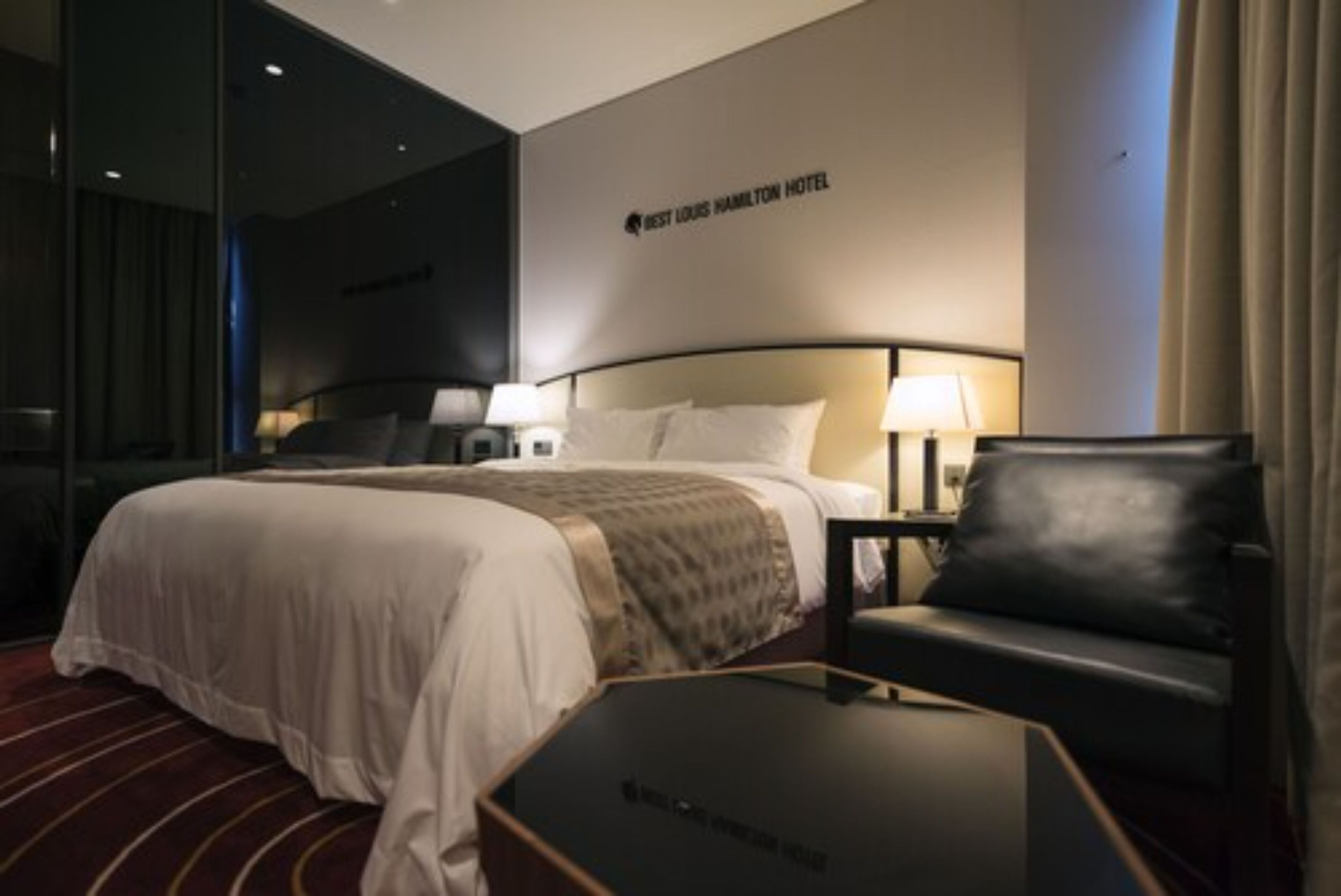 Een hotelkamer met een groot bed, twee nachtkastjes met lampen, een zwarte leren fauteuil en een glazen scheidingswand.