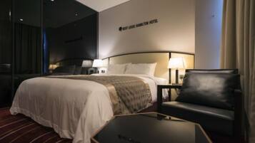 Een hotelkamer met een groot bed, twee nachtkastjes met lampen, een zwarte leren fauteuil en een glazen scheidingswand.