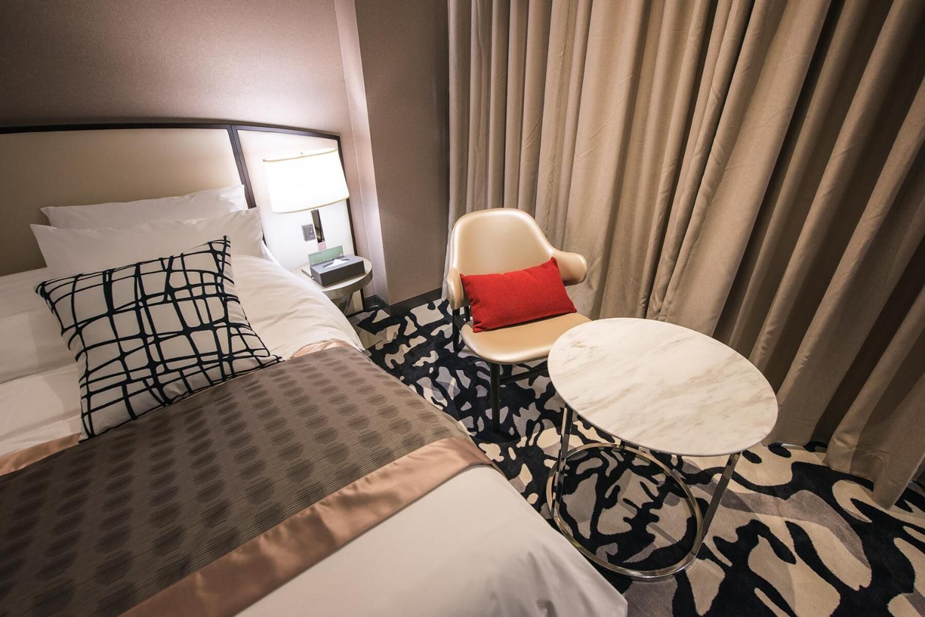 Una habitación de hotel con una cama, una silla, una mesita y una mesita de noche con lámpara.