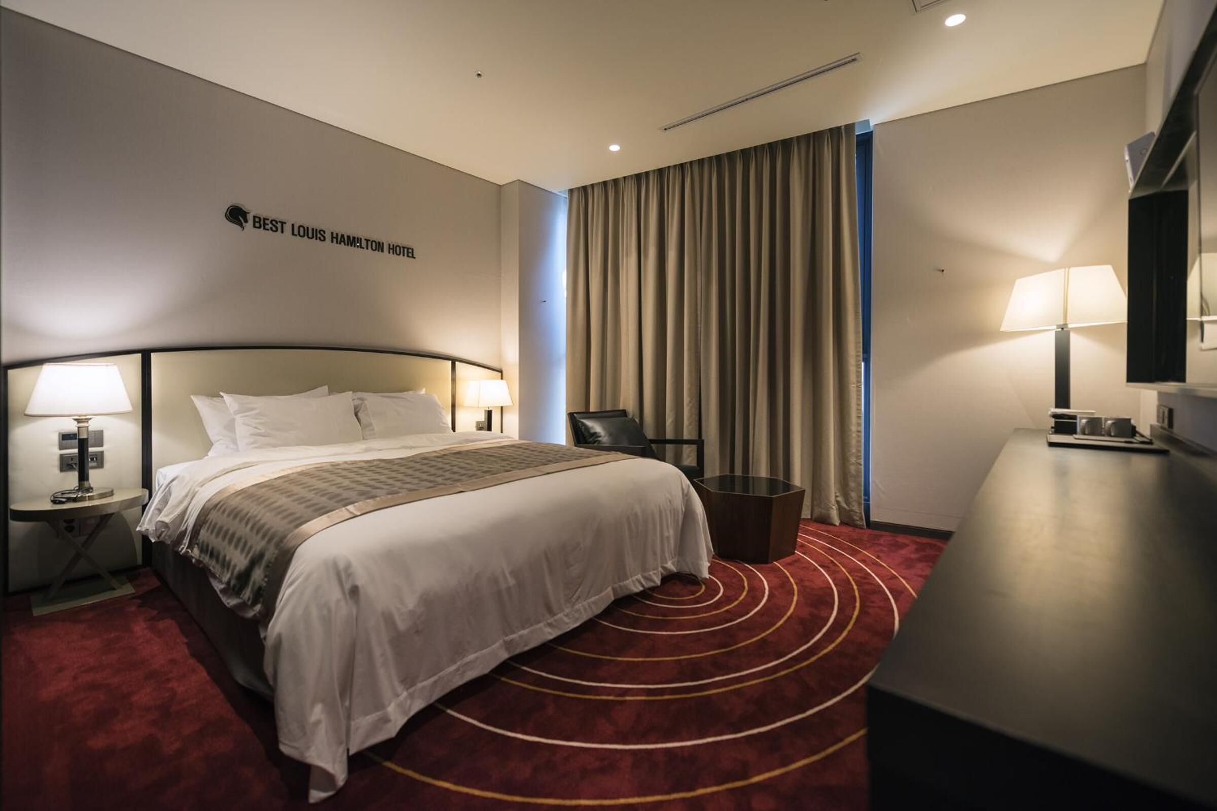 A hotel room with a bed, a desk, a chair, a lamp, and a sign on the wall.