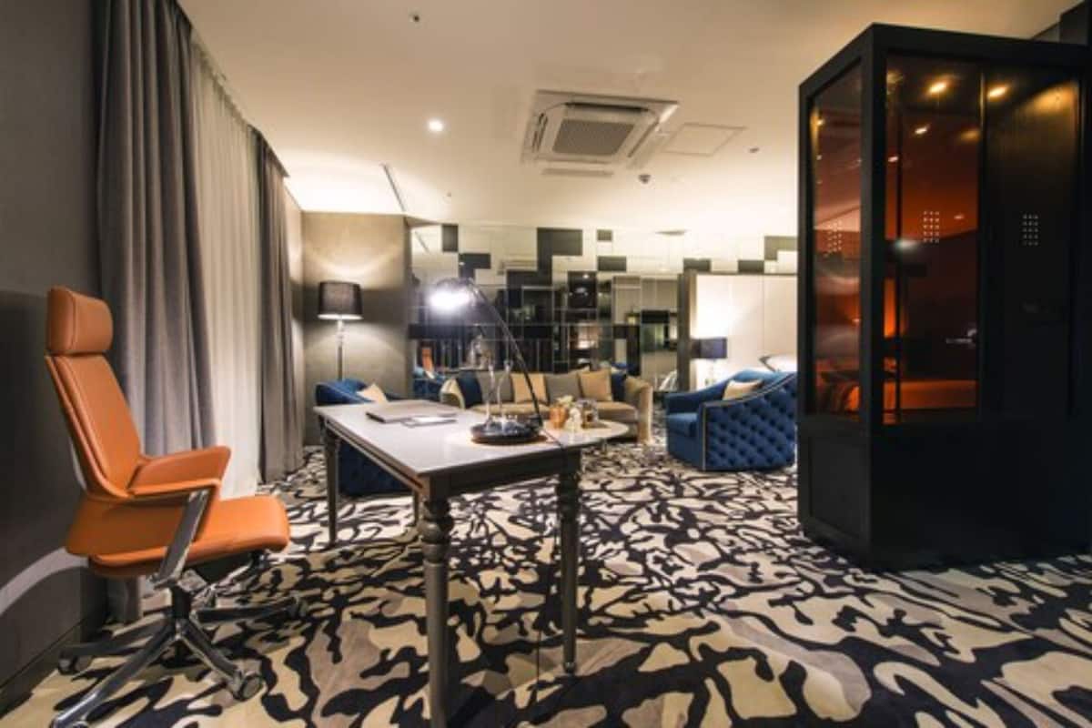 A hotel room with a modern design, featuring a patterned carpet, a dining table with chairs, a sofa, and a separate seating area with a lamp.