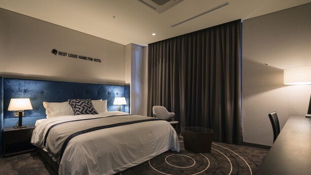 A modern hotel room with a large bed, two bedside tables with lamps, a chair, a small table, and a large window with curtains. - Best Louis Hamilton Hotel Changwon (Changwon)
