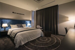 Una habitación de hotel moderna con una cama grande, dos mesitas de noche con lámparas, una silla, una pequeña mesa y un ventanal con cortinas.