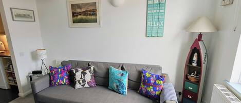 A living room with a grey sofa, colorful cushions, a framed landscape painting, a wall-mounted light, and a decorative item resembling a lighthouse.