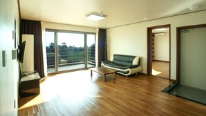 A spacious living room with a wooden floor, a sofa, a coffee table, and a sliding glass door leading to a balcony. - Oasis Pension (Seogwipo)