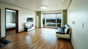 A spacious hotel room with a wooden floor, a sofa, a TV, and a balcony with a view. - Oasis Pension (Seogwipo)