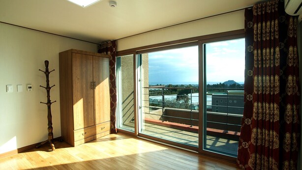 A room with a wooden wardrobe, a coat rack, and a large sliding glass door leading to a balcony with a view of buildings and a mountain. - Oasis Pension (Seogwipo)