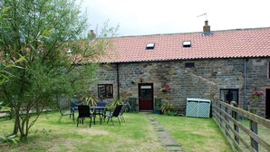 A stone building with a red-tiled roof, a wooden fence, and a paved pathway leading to a table and chairs in the yard.