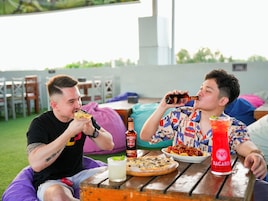 Dos personas sentadas en una mesa al aire libre, con comida y bebidas, rodeadas de sillas y un césped.