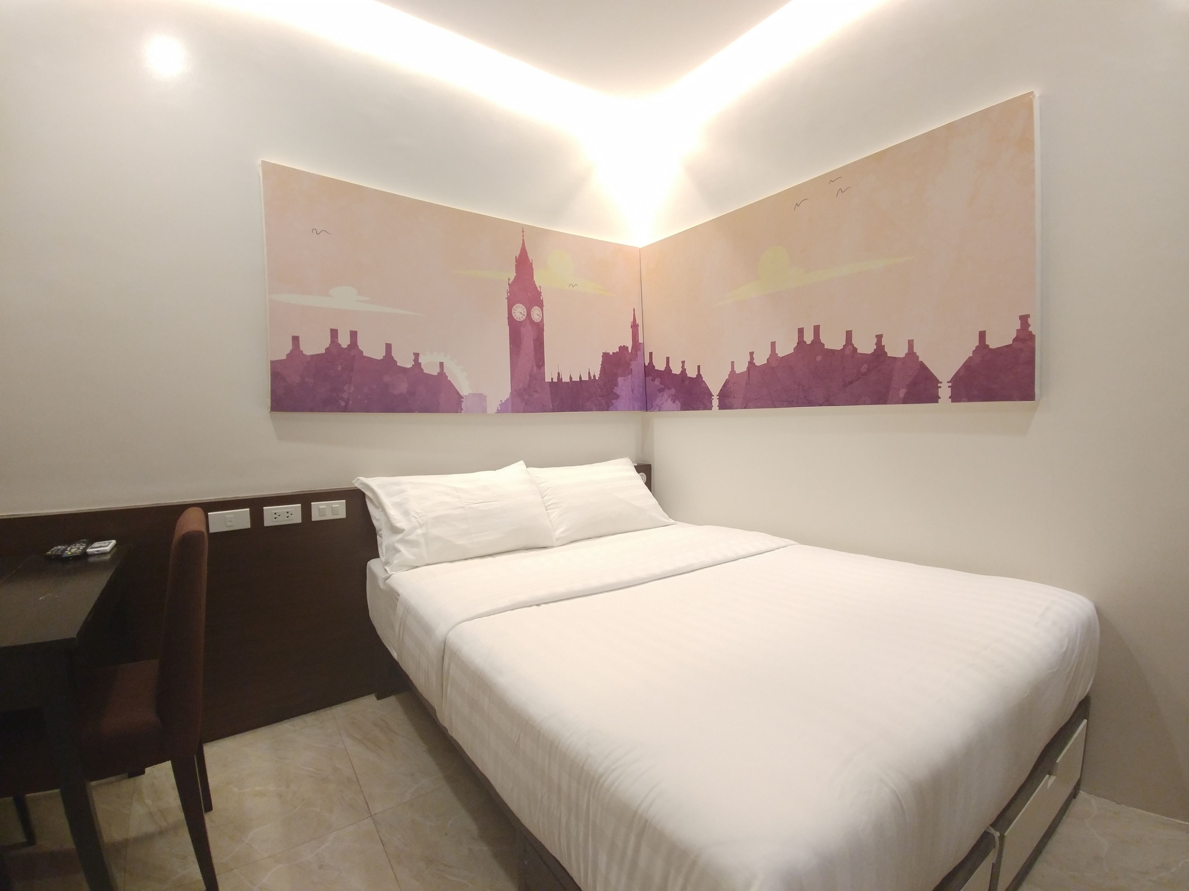 A hotel room with a bed, a desk, and a chair. There is a wall-mounted artwork depicting a city skyline.