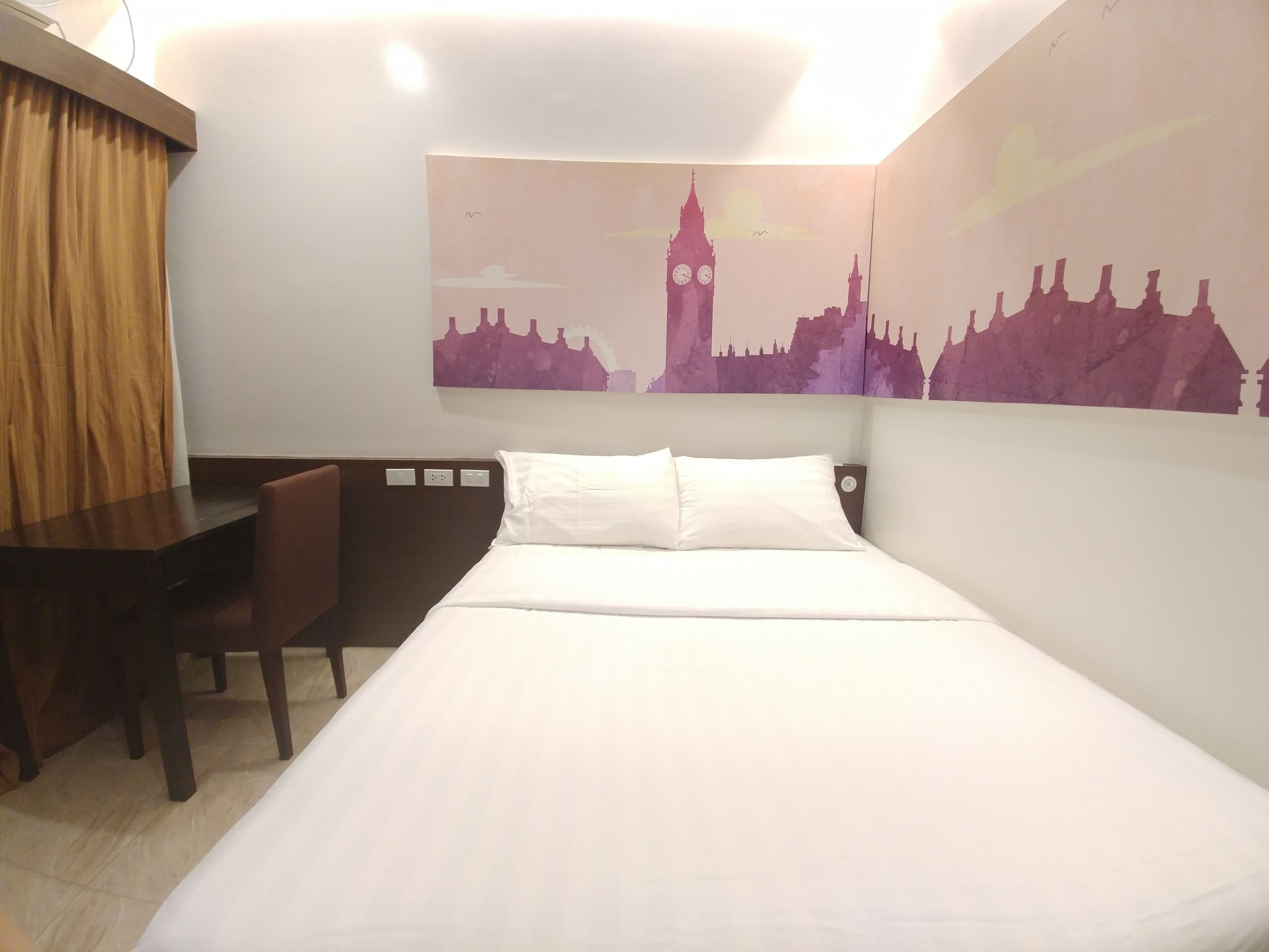 Habitación de hotel con cama, escritorio y silla. Un gran mural en la pared muestra el horizonte de una ciudad.