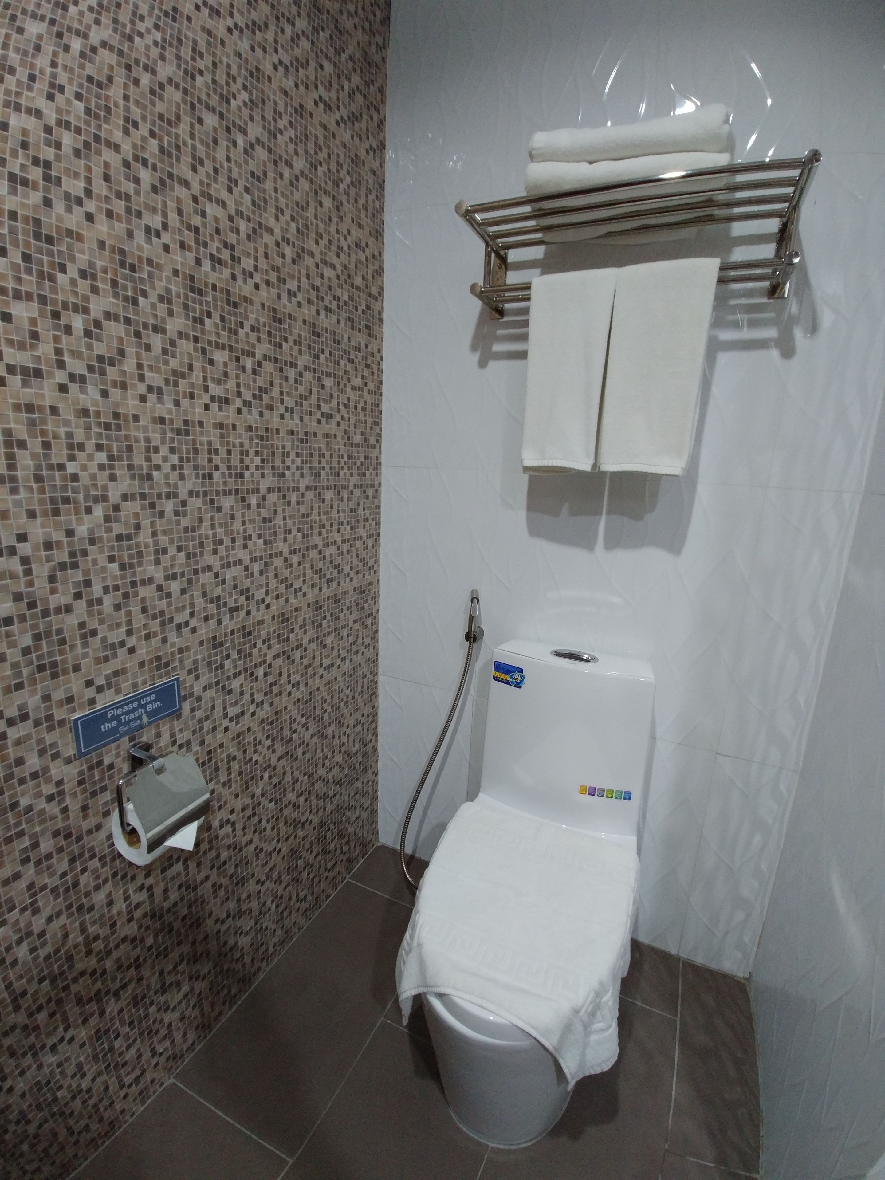 A bathroom with a mosaic tile wall, a toilet with a paper towel dispenser, a towel rack with towels, and a sign instructing to use the trash bin.