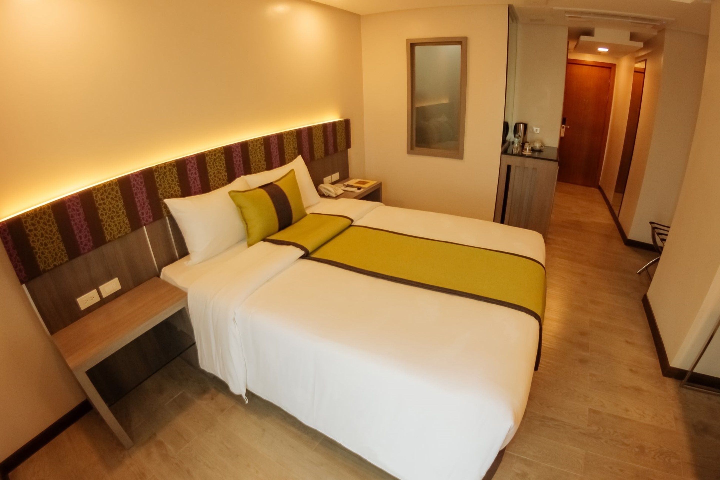 A hotel room with a large bed, bedside table, and a mirror. There is a desk with a chair and a small kitchenette in the corner.