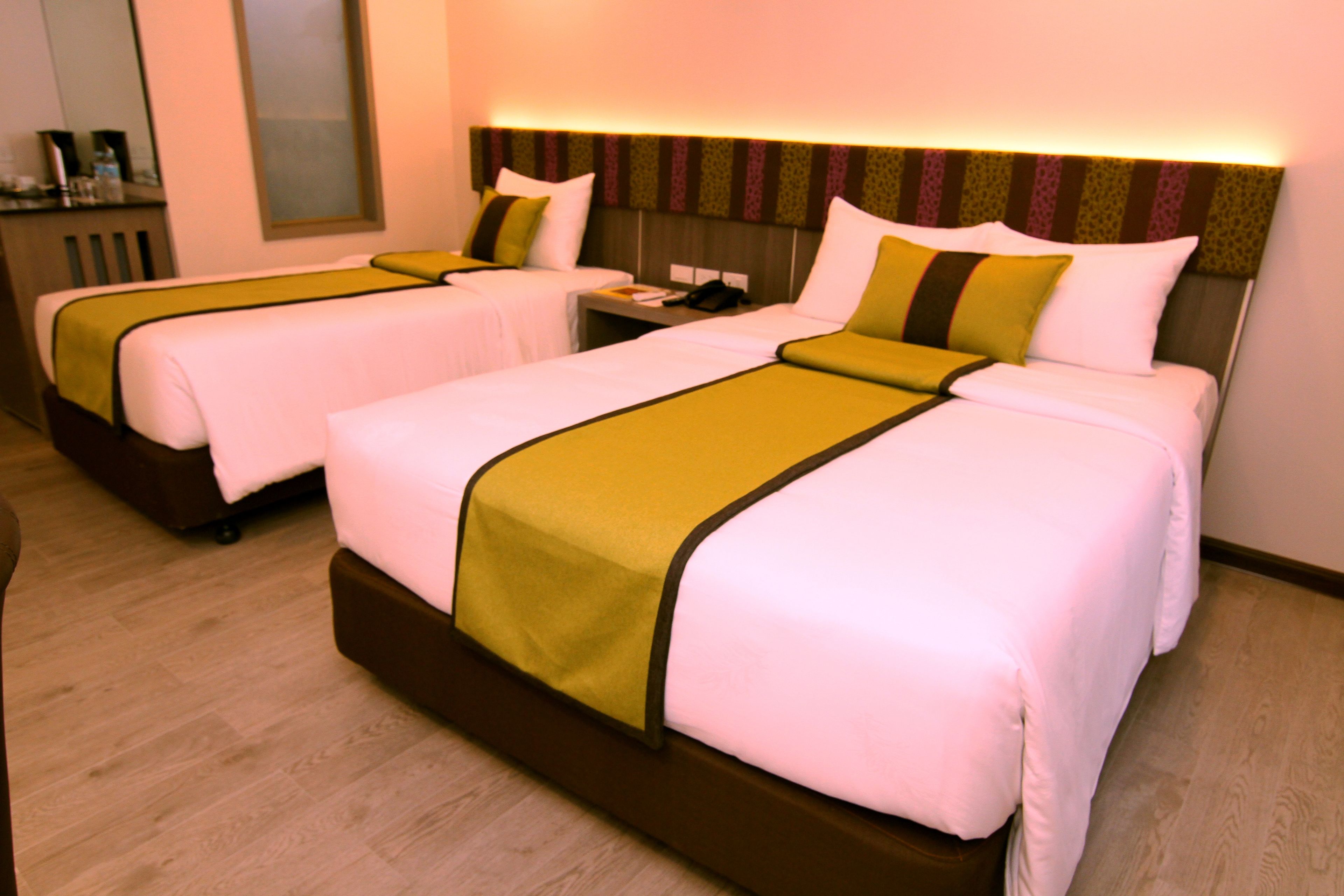 A hotel room with two beds, each with a white and green bedspread, a wooden headboard, and a nightstand with a lamp.