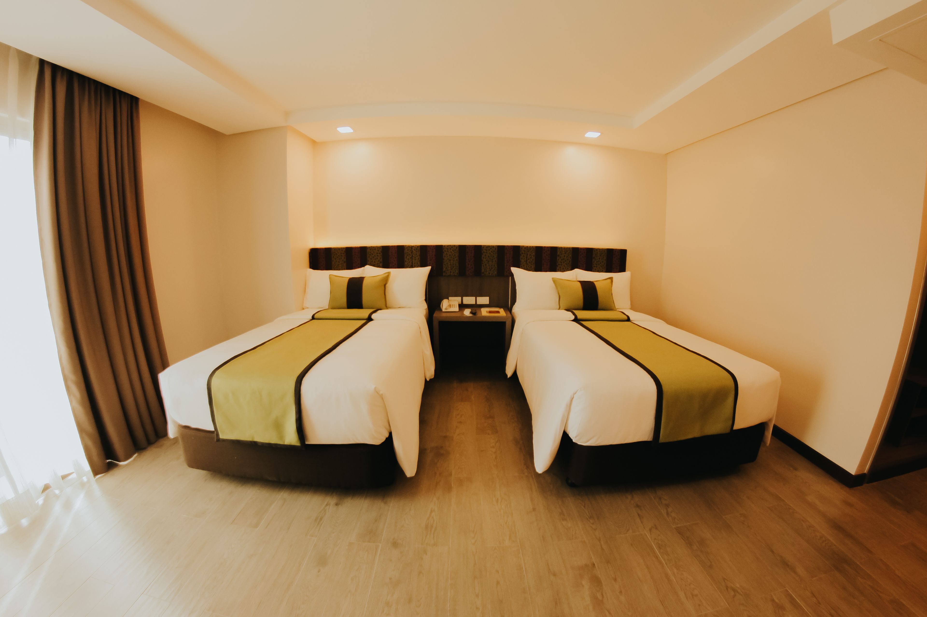 Une chambre d’hôtel avec deux lits simples, une grande fenêtre avec des rideaux, un plancher en bois et une tête de lit sombre.