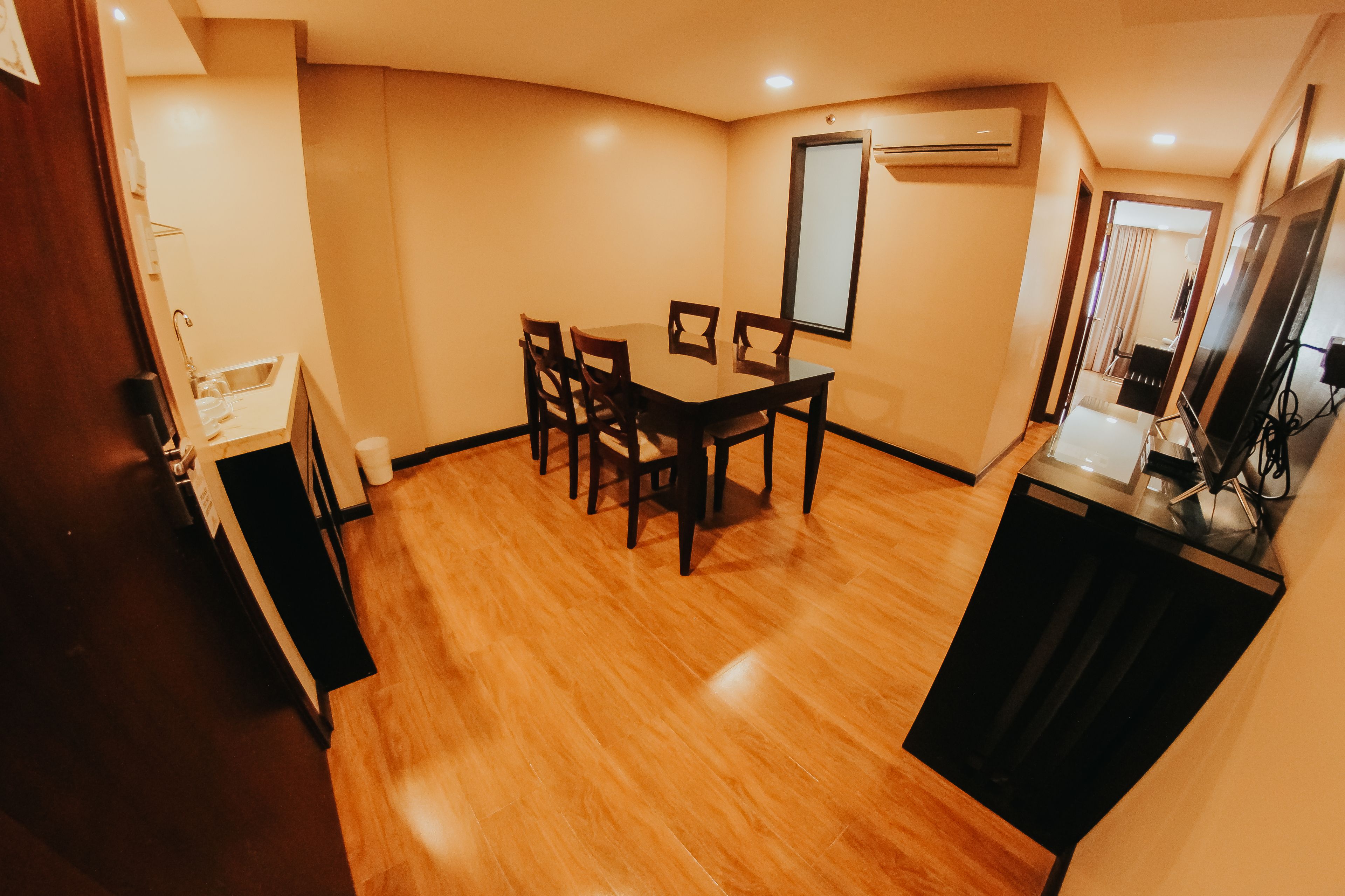 A compact hotel room with a wooden floor, a dining table with chairs, a small kitchenette, and a large mirror.