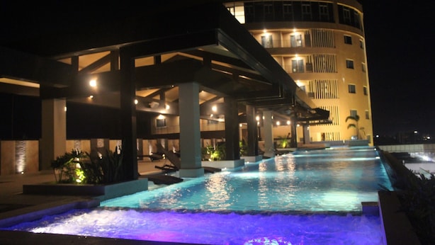 Vista nocturna de un hotel con piscina, mobiliario exterior y un edificio de varios pisos con balcones.