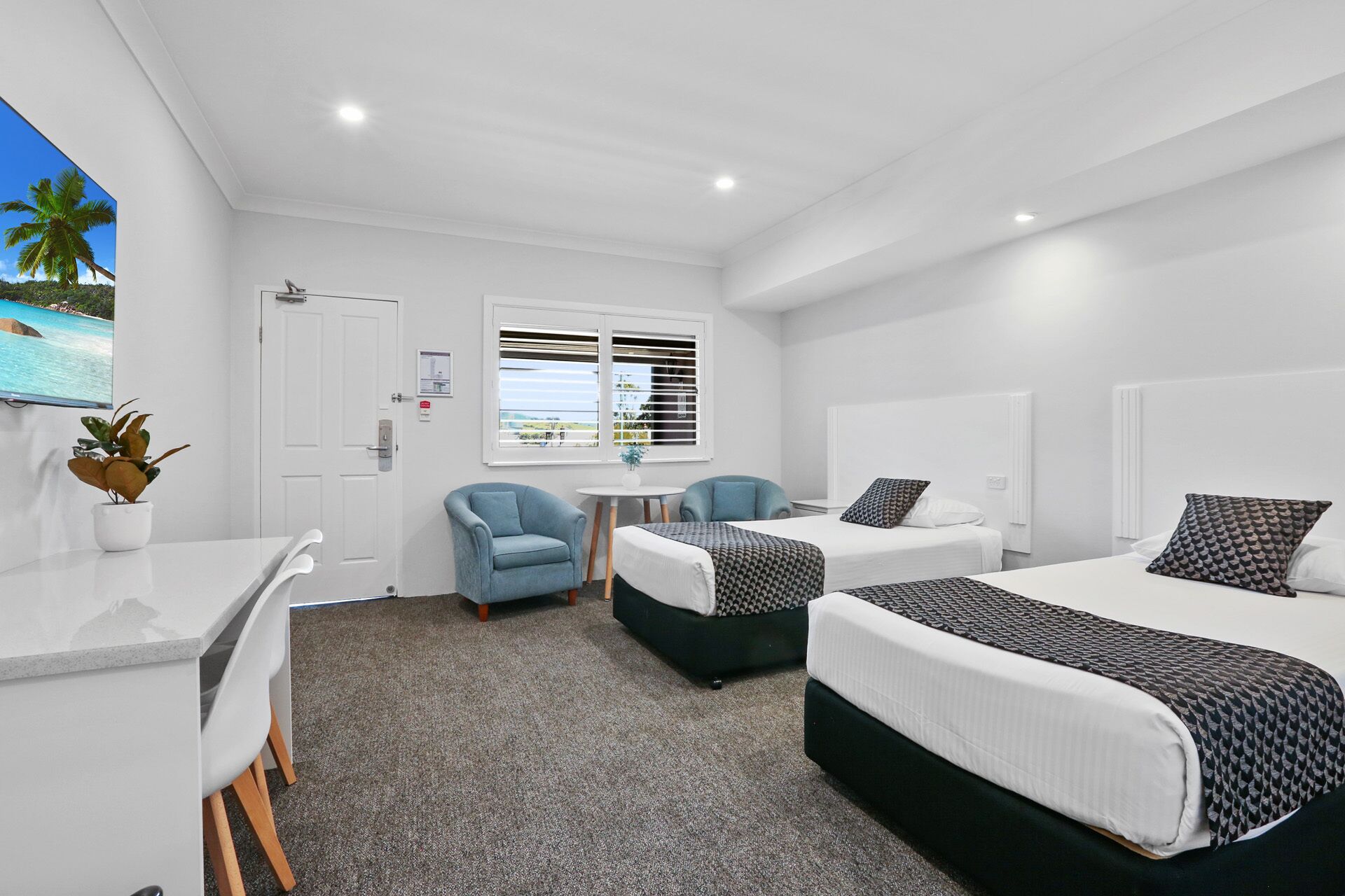 A hotel room with two beds, a desk, a chair, a small table, and a window with blinds.