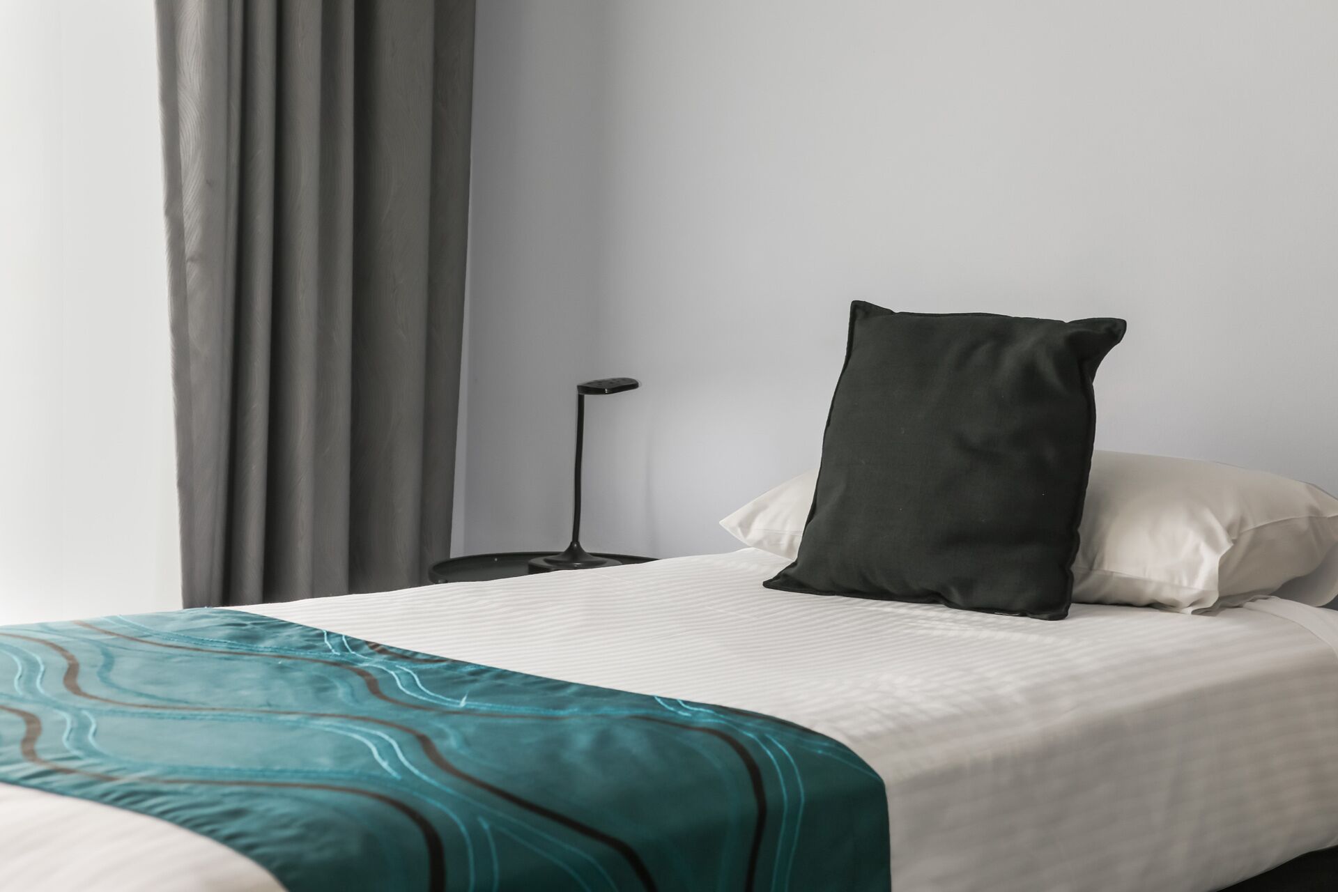 A neatly made bed with a teal patterned blanket, a black pillow, and a white pillow.
