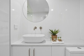 A modern bathroom with a white sink, two faucets, a round mirror, and a small potted plant on the countertop.