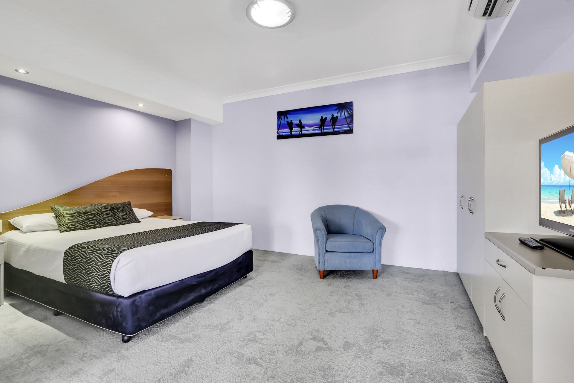 A modern hotel room with a bed, a chair, a TV, and a painting on the wall.